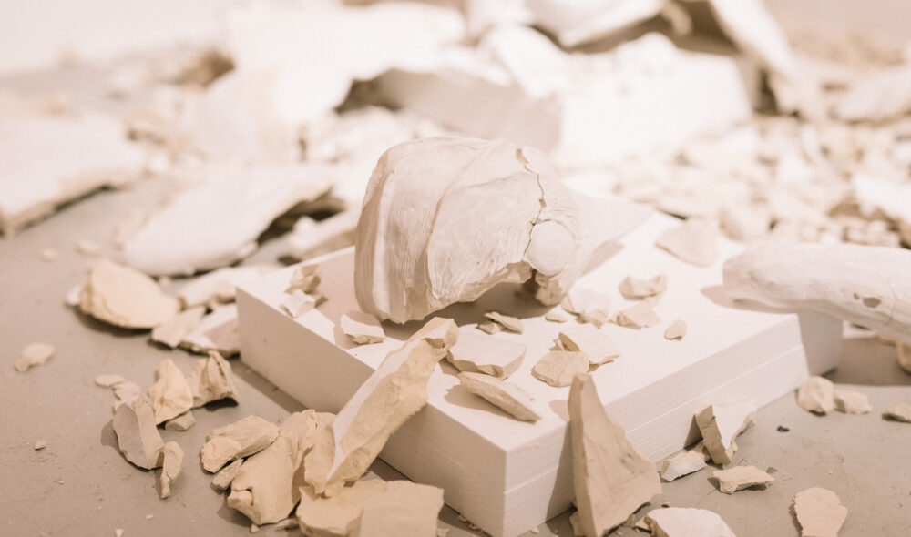 A large, broken ceramic bird’s head sits on a low plinth surrounded by smashed pieces of white clay.