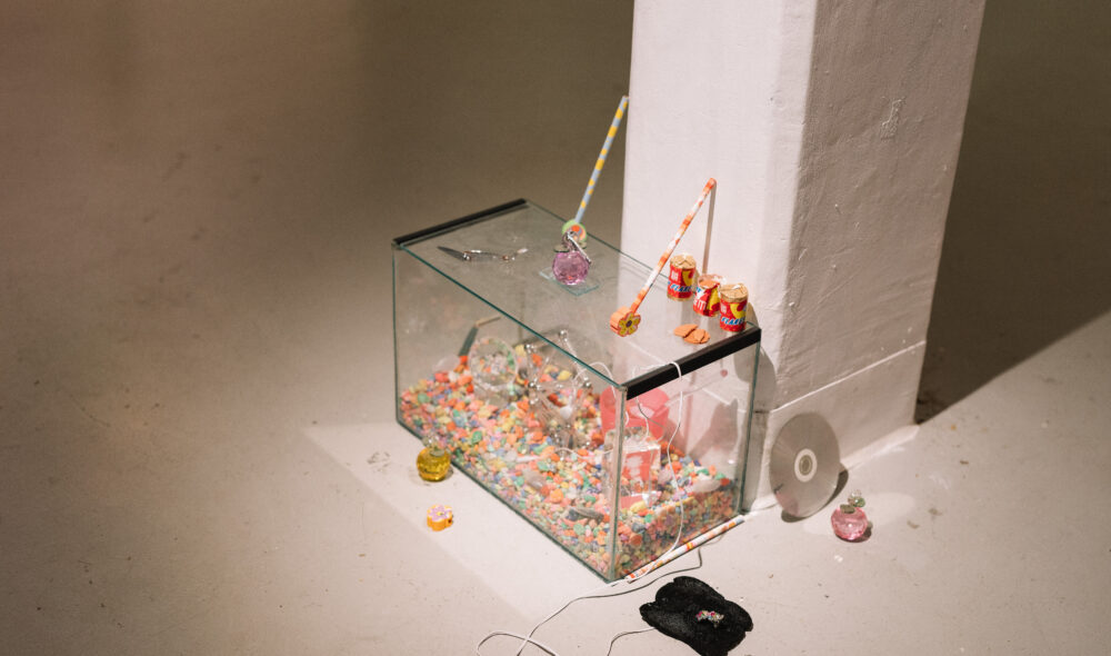 A fish tank sits at the base of a white column in the Nexus Gallery. Empty of water, it is instead full of coloured gravel, clear glass trophies, and small trinkets like toys, erasers and white headphones.