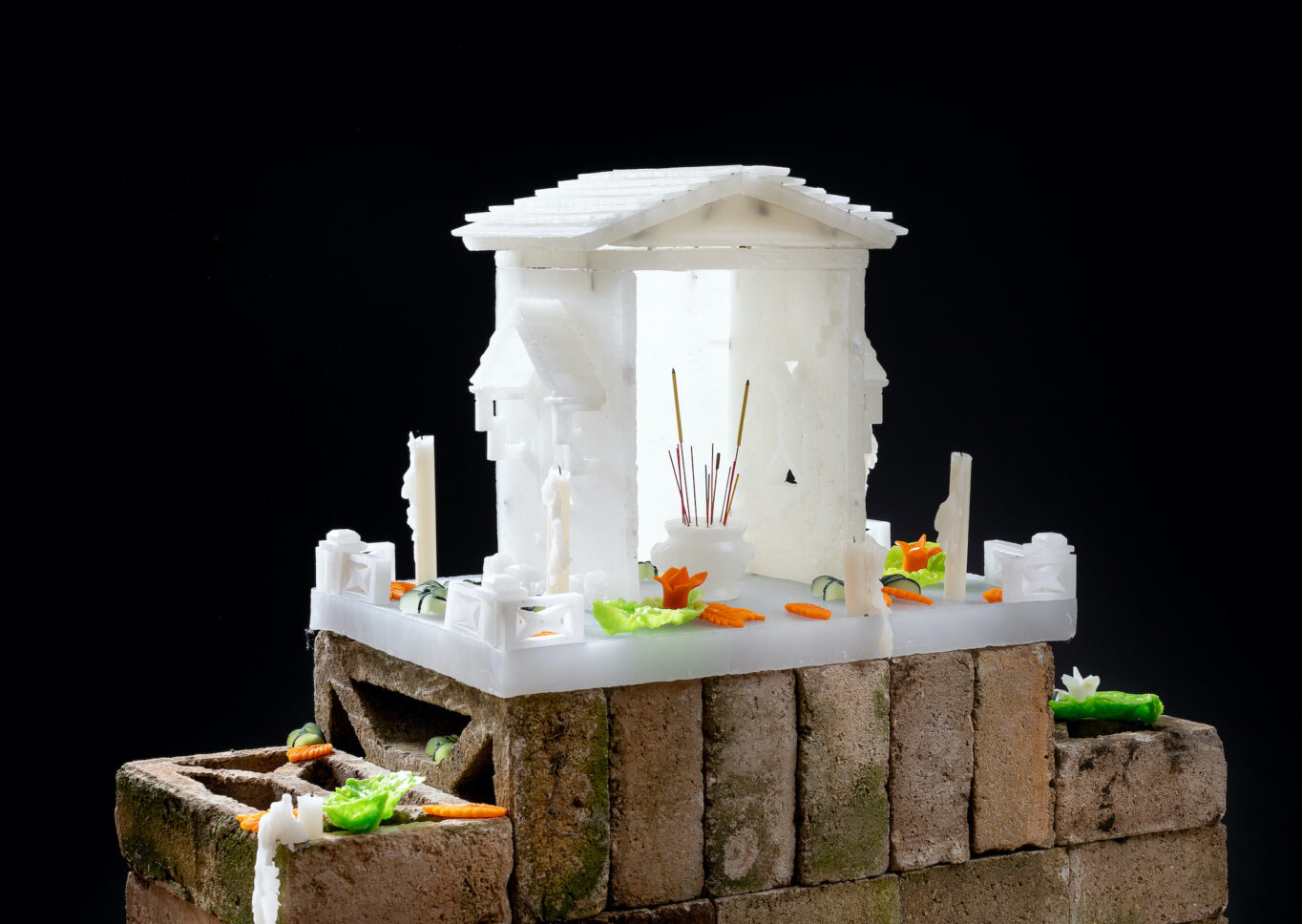 A small white shrine consisting of two side walls and a peaked roof rests on a stack of brown bricks. Within the shrine is a small white vase holding incense sticks, and displayed around it are white candles and small pieces of finely cut orange and green vegetables.