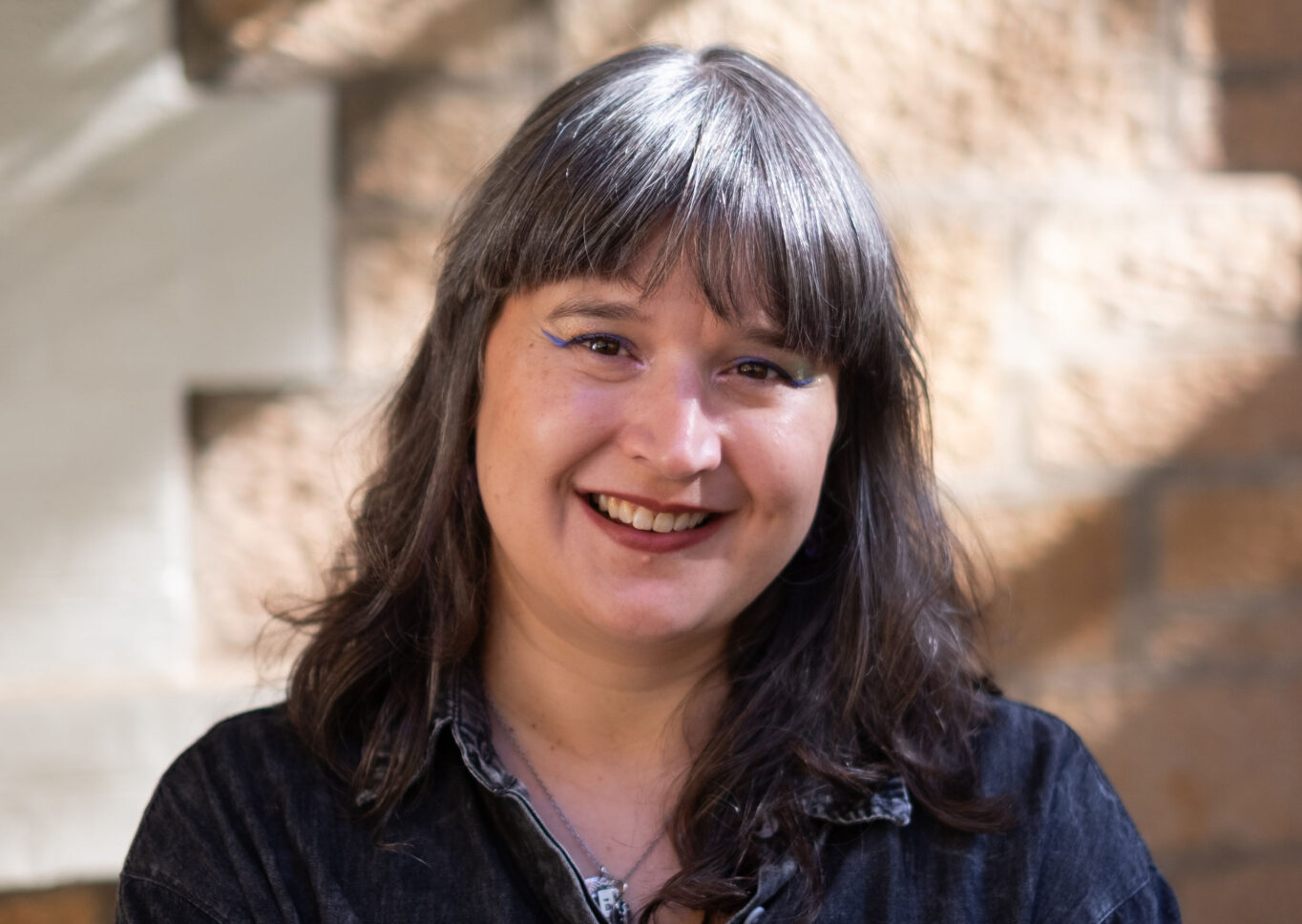 Woman of European and Assyrian ancestry in her mid-40s with long salt and pepper hair smiles broadly at the camera.  She is wearing a black long sleeve shirt in front of a sandstone building. Photo by Kathryn Harvy.