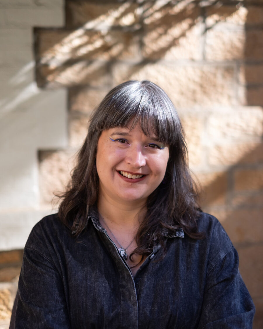 Woman of European and Assyrian ancestry in her mid-40s with long salt and pepper hair smiles broadly at the camera.  She is wearing a black long sleeve shirt in front of a sandstone building. Photo by Kathryn Harvy.