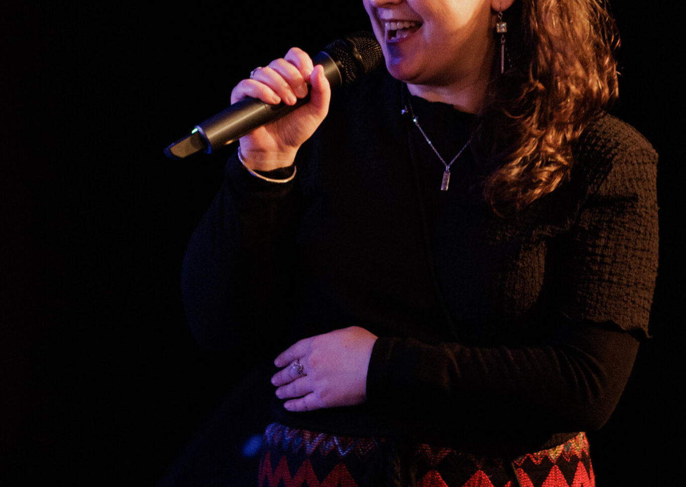 Woman of European and Assyrian ancestry in her mid-40s with long salt and pepper hair smiles broadly while talking into a microphone.  She is wearing a 1970s black and red woven skirt with a zig zag pattern and black tee. Photo by Bri Hammond.