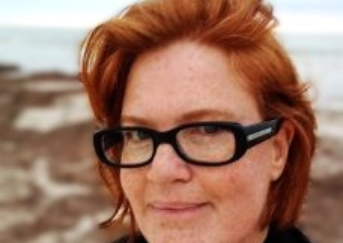 A picture of Michele Saint-Yves, a woman with soft wave thick red hair to top of shoulders. Eye colour: Blue-Grey Accessories: Black rectangular glasses Other: Covered in freckles