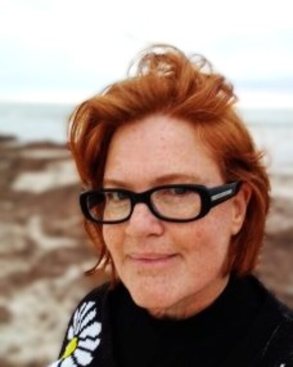 A picture of Michele Saint-Yves, a woman with soft wave thick red hair to top of shoulders. Eye colour: Blue-Grey Accessories: Black rectangular glasses Other: Covered in freckles